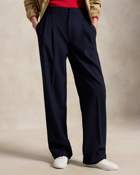 Person wearing navy pleated pants with a beige jacket and white shoes on a neutral background