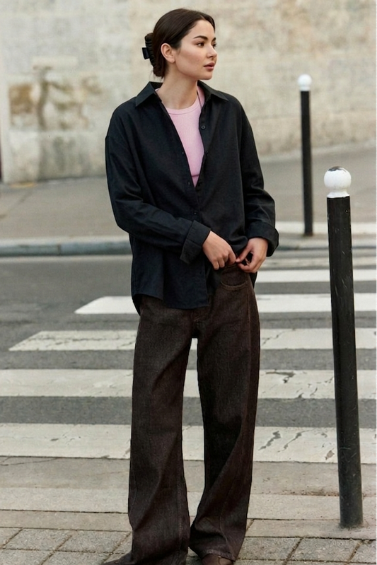 Hania amir standing on a street corner wearing a black button down shirt and brown pants.